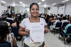 Descrição da imagem. Foto horizontal tirada do fundo de um auditório lotado de pessoas sentadas , mostra em primeiro plano uma mulher em pé, sorrindo, segurando à altura do peito um certificado com logomarca no canto superior esquerdo do Senac.  Fim da descrição.