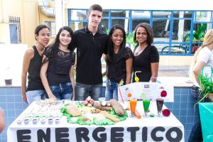 descrição da imagem. Foto horizontal de um grupo de 5 jovens, em pé, lado a lado, olhando para a frente e sorrindo, vestidos em calça jeans e camisas pretas, sendo um rapaz ao meio e duas moças em seu lado direito e mais duas em seu lado esquerdo.  À frente deles, uma mesa retangular coberta com um tecido branco e à sua frente, colada a palavra Energético em letras maiúsculas pretas. Sobre a mesa, da esquerda para a direita, copinhos descartáveis tipo de cafezinho formam uma fila na horizontal com alguns grãos dentro, ao centro da mesa, folhas de alface formam um círculo e sobre as mesmas, raízes, grãos e pão integral fatiado, e à direita, três taças de vidro transparente com um suco e o alimento que o representa ao lado, sendo o primeiro de cenoura, o segundo de limão e o terceiro de beterraba. Fim da descrição.