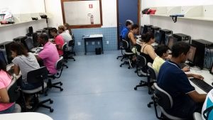 Descrição da imagem. Foto horizontal de um laboratório de informática de piso azul e paredes brancas, sendo que nas paredes da esquerda e da direita aparecem 6 pessoas sentadas à frente de computadores arrumados lado a lado sobre mesas também organizadas lado a lado.  Acima da altura das pessoas, há prateleiras brancas com algumas pastas e mochilas. Na parede ao fundo, uma grande janela de vidro e do lado direito uma porta azul. Fim da descrição.