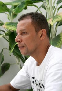 Descrição da imagem. Foto vertical de um homem, pele moreno clara, de perfil para a esquerda, com olhar voltado para esse lado, camisa branca e ao fundo, folhas largas e verdes de uma planta e parede branca mais ao fundo. Fim da descrição.