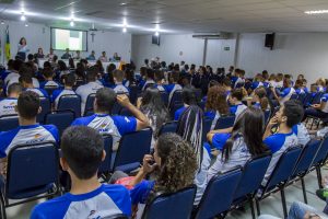 Descrição da imagem. Foto horizontal com perspectiva do fundo de um auditório de cadeiras azuis, lotado de jovens com camisa do Senac, e à frente deles, bancada branca com pessoas sentadas à mesma e telão ao fundo. Fim da descrição.