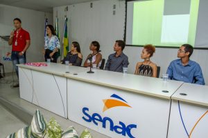 Descrição da imagem. Foto horizontal de uma bancada branca com logo do Senac na frente, pessoas sentadas atrás da mesma e telão ao fundo. Do lado esquerdo da bancada, de pé, um rapaz com camisa vermelha e calça jeans, segura microfone próximo à boca. Fim da descrição.