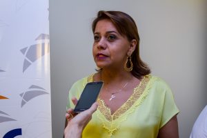 Descrição da imagem. Foto horizontal de meio busto de uma mulher de semiperfil para a esquerda, com cabelos castanhos e curtos, blusa amarela, com boca semiaberta. À sua frente, uma mão segura um celular à altura próxima de seu queixo. Fundo branco. Fim da descrição.