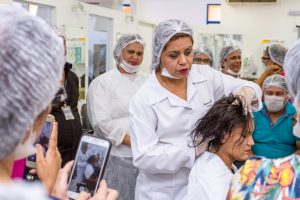 Descrição da imagem. Foto horizontal de uma mulher com touca e jaleco brancos segurando os cabelos de uma mulher sentada, com roupa branca e olhos fechados. Ao fundo,grupo de  pessoas usando touca, alguns observam a mulher em destaque outros sorriem e se olham. No lado esquerdo, aparece parcialmente uma moça de costas segurando o celular à frente da mulher com a câmera ativada. Fim da descrição. 