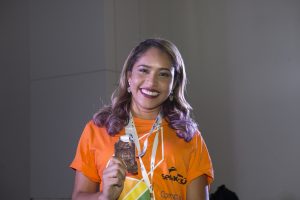 Descrição da imagem. Foto horizontal de busto para cima de uma jovem de cabelos claros, cacheados nas pontas, olhando para a frente e sorrindo, camisa laranja com logomarca do Senac, cordão pendurado no pescoço com medalha na ponta, a qual está segura por sua mão direita. Fundo com parede branca. Fim da descrição. 