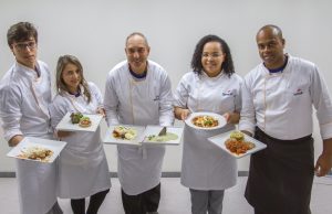 Descrição da imagem. Foto horizontal de cinco pessoas de pé, de frente, olhando para a câmera, vestindo avental branco de cozinheiro com logomarca do Senac