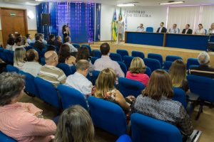 Descrição da imagem. Foto horizontal do fundo de um auditório de poltronas azuis, com muitas pessoas sentadas e duas filas de poltronas vazias na frente. No palco do auditório, cerca de um metro acima do nível das poltronas, bancada de cor azul na parte frontal, com seis pessoas sentadas à mesma. Ao fundo, do lado esquerdo, três bandeiras em mastro, duas verde e amarelas e uma branca, e na parede ao fundo letras formam AUDITÓRIO OVIÊDO TEIXEIRA, do lado direito, persianas brancas. Fim da descrição.