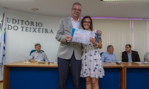 Descrição da imagem. Foto horizontal parcial de um homem e uma mulher lado a lado, juntos, de pé, sorridentes, ele do lado esquerdo e ela do lado direito. A mulher, mais baixa, usa vestido claro estampado na altura do joelho, tem pele moreno clara, cabelos curtos e lisos, óculos de grau, a mão direita por trás do homem e a mão esquerda segura um certificado juntamente com o homem, que usa a mão direita. Ele, mais alto, tem bigode e cabelos grizalhos, óculos de grau, terno cinza e camisa branca, e calça escura. Ao fundo, bancada de madeira com detalhe azul na parte frontal, homens com camisa social sentados à mesma. Do lado esquerdo, mastro com uma bandeira de cor predominante branca, na parede ao fundo de cor branca, letras formam o nome AUDITÓRIO, e logo abaixo, o nome do mesmo que não aparece por completo, e do lado direito, persianas brancas. Fim da descrição.