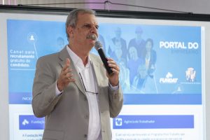 Descrição da imagem. Foto horizontal parcial de um homem de pé, em semiperfil para a direita, bigode e cabelos grizalhos, terno cinza e camisa branca, óculos de grau pendurados por cordão no pescoço, segura com a mão esquerda um microfone próximo à boca, e com a mão direita gesticula. Ao fundo, tela com projeção de imagens e textos. Fim da descrição.