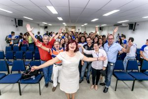 Foto horizontal de dezenas de pessoas em pé no corredor central do auditório do Senac. Todas estão olhando para a frente, sorridentes, com os braços levantados. À frente, no meio, está a palestrante, logo atrás, estão os instrutores e os alunos.