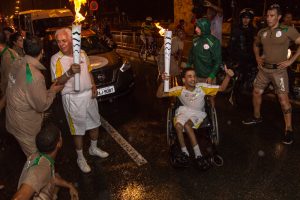 Foto horizontal, à noite, de dois condutores da tocha olímpica na avenida Ivo do Prado. Ao centro, duas pessoas seguram, cada uma, uma réplica da tocha olímpica acesa, à esquerda, um senhor parcialmente calvo de cabelos totalmente brancos, olha para uma pessoa que está na lateral da avenida, o outro, do lado direito é um jovem cadeirante, sorridente, faz sinal de positivo com uma das mãos. Ambos vestem uniforme branco, composto por bermuda e camisa com detalhes amarelos, e, na altura do peito pintado em dourado o nome RIO 2016 e o símbolo das olimpíadas. Ao fundo dos dois, um carro de polícia. Nas laterais da avenida, barreira de pessoas com uniforme cinza com detalhes verde nos ombros e nome RIO 2016 pintado de branco na perna esquerda da bermuda e nas costas da camisa e dos casacos.