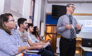 Descrição da imagem. Foto horizontal de parte de uma sala de aula, à esquerda, quatro pessoas, dois homens e duas mulheres, sentados em carteiras com pasta do Senac sobre as mesmas, organizados em fila, lado a lado, olham para um homem que está à sua frente, em pé, roupa social e gravata, usa óculos, cabelos grisalhos, gesticula e olha para a direita da imagem com a boca entreaberta. Ao fundo, paredes brancas e tela projetada ao fundo da sala. Fim da descrição.