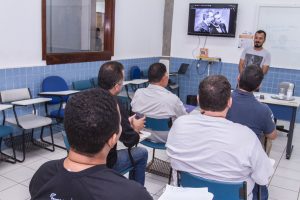 Descrição da imagem. Foto horizontal do fundo de uma sala de aula, mostra de costas cinco homens sentados em carteiras organizadas em duas filas ao centro e à frente deles um homem em pé, atrás do birô, olha para os mesmos. Ao fundo, televisão presa à parede no canto esquerdo superior, as paredes são da metade para baixo de pequenos azulejos azuis e a outra metade pintada de branco. Fim da descrição.