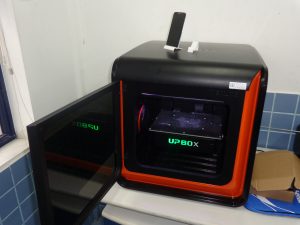 Descrição da imagem. Foto horizontal de uma impressora 3 d Up Box sobre uma pequena mesa branca. A impressora é de cor preta, formato cúbido, com pouco mais de meio metro de largura, de altura e de profundidade. Sua face lateral é uma porta de vidro, a qual está aberta para o lado esquerdo. Em seu interior, uma bandeja suspensa e na frente escrito UP BOX com letras maiúsculas na cor verde. Sobre a impressora, estão duas pequenas peças brancas com uma base e uma lateral apenas, inclinada para trás. Sobre uma delas, um celular na posição vertical. Fim da descrição.