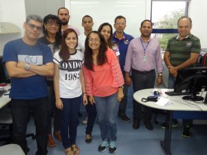 Descrição da imagem. Foto horizontal de um grupo de pessoas em pé, numa sala de aula, olhando para a frente. À esquerda, oito alunos, homens e mulheres, juntos, com um leve sorriso. À direita, de camisa social lilás, calça jens e crachá do Senac, o coordenador de informática Cláudio, e de camisa verde e calça jeans o professor da turma, Francisco. À frente dos dois, um monitor, mouse e teclado pretos sobre uma mesa branca. Fim da descrição.
