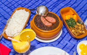 Descrição da imagem. Foto horizontal com vista superior de três recipientes de barro com alimentos preparados. Da esquerda para a direita, uma travessa com arroz branco e uma colher metálica, no meio uma tigela redonda com feijão preto cozido e uma linguiça cortada ao meio sobre o mesmo e uma concha metálica apoiada na borda, e à direita, uma travessa com abóbora cortada em cubos, couve cortada em tiras e alguns pedaços de torresmo e um garfo metálico. Na parte inferior, um copo de vidro com suco de laranja e decorado com uma fatia da fruta presa à borda e canudo de plástico. Parte de outros pratos na parte inferior. Fim da descrição. 
