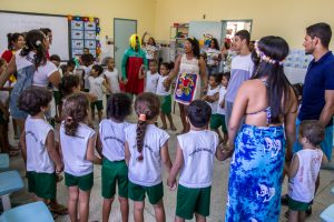 Foto horizontal de uma sala de aula sem as carteiras, no centro um círculo de pessoas em pé, formado por jovens e crianças de cerca de 5 anos. As crianças estão vestidas com um fardamento de bermuda verde e camisa regata branca com o termo Educação Infantil escrito na cor preta nas costas escrito. Todos estão sorrindo e de mãos dadas. As paredes da sala estão decoradas com flores de papel, quadro branco e prateleiras com cadernos coloridos. Fim da descrição.