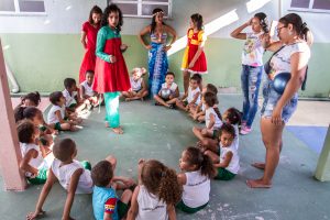 Foto horizontal de um grupo de crianças sentadas em um chão de cimento, formando um círculo. Ao centro, em pé, uma jovem com vestido de manga longa verde e vermelho olha para as crianças e gesticula com os braços. Fora do círculo, em pé, olhando para as crianças, outras jovens, duas com a camisa branca com o nome folclore, uma com o vestido amarelo e vermelho, outra com a coroa de flores na cabeça, veste parte superior do biquíne e saída de praia amarrada à cintura azul e a última com vestido longo vermelho. Fim da descrição. 