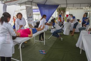 Descrição de imagem. Foto horizontal de pessoas sob tendas brancas deitadas em macas e/ou sentadas em cadeiras de massagem e outras pessoas de pé, com farda do Senac fazendo massagem nas mesmas. Chão tipo carpete verde. Fim da descrição.