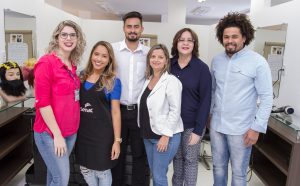 Descrição da imagem. Foto horizontal de seis pessoas adultas em pé num salão de beleza de paredes brancas e balcões azuis. Da esquerda para a direita, uma mulher de calça jeans e blusa rosa, cabelo loiro cacheado, óculos de grau e crachá do Senac, ao lado, mulher mais baixa, blusa azul e calça jeans, avental preto até o joelho, ao lado, homem alto, cabelo curto, cavanhaque, camisa social branca e calça preta, ao lado, mulher baixa, blusa preta, calça jeans e blaser branco, cabelo loiro e liso, ao lado mulher de cabelos castanhos e lisos, óculos de grau, camisa de manga comprida azul escura e calça cinza, e por último, homem de cabelos cacheados e volumosos, e cavanhaque castanhos, camisa social azul escura e calça jeans desbotada. Fim da descrição.