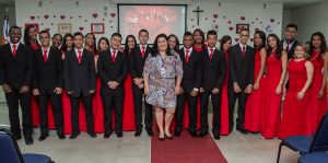 Descrição da imagem. Foto horizontal de um grupo de cerca de 25 pessoas jovens em pé, posando para a foto sorrindo. Ao fundo estão moças com vestidos longos vermelhos, todos do mesmo modelo, na fila da frente, rapazes de terno, calça social e sapato pretos, camisa branca e gravata vermelha. Mais à frente, mulher com vestido estampado na altura do joelho, pele clara e cabelos pretos e lisos e sapato preto. Ao fundo, parede branca decorada com corações pequenos, vermelhos, e ao centro um telão com a imagem de um coração projetada. O piso é branco e um tapete comprido vermelho ao centro. Nos cantos inferiores da imagem, parte de cadeiras azuis. Fim da descrição. 