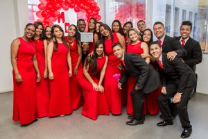 Descrição da imagem. Foto horizontal de um grupo de jovens agrupados em pé sorrindo e posando para a foto. A maioria são moças, que se concentram do meio para o lado esquerdo da imagem, vestem vestido vermelho longo de alças largas, e no canto direito, alguns rapazes, vestem terno, calça e sapatos pretos, camisa social branca e gravata vermelha. Ao fundo, parede branca com janelas de vidro e bexigas vermelhas formando um arco. Fim da descrição.
