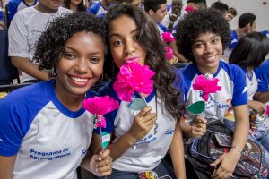 Descrição da imagem. Foto horizontal, parcial, de três moças com farda do Senac sentadas num auditório. com várias outras pessoas fardadas também. As jovens tem cabelo cacheado, sendo as das extremidades o cabelo curto e a do meio o cabelo comprido, seguram uma flor de papel rosa com uma folhinha verde no cabo. Fim da descrição.