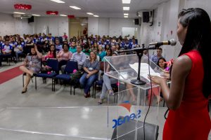 Descrição da imagem.  Foto horizontal de uma jovem de vestido vermelho de costas, com as mãos sobre um púlpito transparente com o nome do Senac e próxima a um microfone, olhando para a plateia em um auditório. A plateia está repleta de jovens com a farda do Senac e as duas filas da frente estão ocupadas por pessoas com crachá do Senac. O espaço tem paredes, teto e piso claros e um tapete vermelho no corredor central. Fim da descrição.