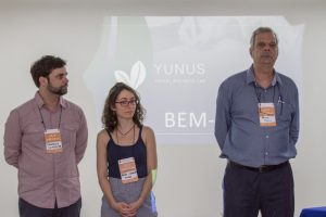 Descrição da imagem. Foto horizontal de três pessoas de pé, de frente, usando crachá laranja. Da esquerda para a direita, um homem de estatura média, cabelos ondulados castanhos e barba, veste camisa social manga longa na cor vinho e olha para seu lado esquerdo com os braços para trás. No meio, uma mulher baixa, cabelos castanhos cacheados, óculos de grau, blusa azul de alças e saia cinza, com as mãos unidas para a frente, olha para baixo. No canto direito da imagem, homem alto, cabelos e bigode grisalhos, camisa social azul de mangas longas, óculos de grau pendurados pelo pescoço, com os braços para trás, olha para a frente. Ao fundo, parede branca com projeção pouco nítida. Fim da descrição.