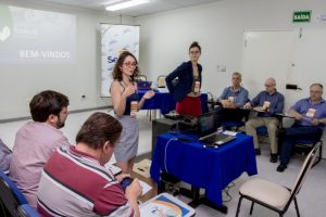 Descrição da imagem. Foto horizontal parcial de uma sala de aula com duas mulheres ao centro, de pé, gesticulando, três homens sentados na parede do fundo da imagem e dois sentados no canto esquerdo inferior, de costas para o ângulo da foto. e de frente para as mulheres. Fim da descrição.