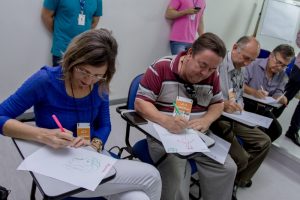 Descrição da foto. Foto horizontal de quatro pessoas com crachá laranja sentadas em carteiras escrevendo em folhas de papel olhando para as mesmas. À esquerda, mulher de cabelos à altura do ombro, levemente loiros, óculos de grau, blusa azul de manga três quartos e calça clara de pernas cruzadas, ao seu lado, homem de cabelos castanhos lisos, penteados para trás, camisa tipo polo de listras horizontais largas em vermelho e bege, óculos escuros pendurados na gola e calça cinza. Ao lado, homem calvo, camisa social cinza e calça marrom, e por último, homem de cabelos grisalhos, óculos de grau, camisa azul e calça preta. Ao fundo, parte de dois homens em pé, encostados na parede. Paredes e piso brancos. Fim da descrição.