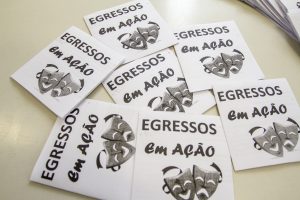 Descrição da imagem. Foto horizontal de vista superior de 7 folhetos de fundo branco dispostos de forma aleatória sobre uma superfície branca e no canto superior da imagem parte de uma uma fileira de folhetos sobrepostos. Os folhetos tem formato retangular na vertical, impressão em preto e branco, na parte superior em caixa alta, EGRESSOS EM AÇÃO. Abaixo, máscaras de drama e comédia que representam o teatro. Fim da descrição.