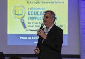 Descrição da imagem. Foto horizontal do senhor Paulo do Eirado de semiperfil para a esquerda, segurando um microfone com a mão esquerda próximo à boca. Ao fundo, parede com projeção de fundo amarelo com letras azuis escrito 1º FÓRUM DE EDUCAÇÃO EMPRESARIAL. Fim da descrição.