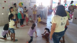 Descrição da imagem. Foto horizontal de um espaço com piso e azulejos brancos, várias crianças, algumas sentadas e outras de pé e as duas moças com roupas coloridas e nariz de palhaço. Fim da descrição.