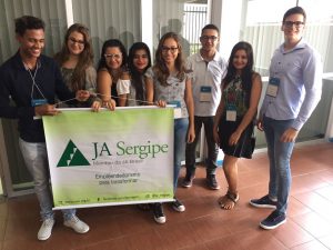 Descrição da imagem. Foto horizontal de um grupo de oito jovens de pé, sorridentes, usando crachá, e os cinco do lado esquerdo seguram juntos um banner branco e verde com o nome JA Sergipe. Fim da descrição.