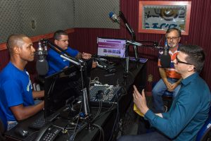 Descrição da imagem. Foto horizontal de quatro homens num estúdio de rádio com bancada e aparelhagem e microfones ao centro. Dois dos homens estão vestidos com camisas azuis e sentam do lado esquerdo da imagem, e olham para os outros dois homens que sentam do lado direito da imagem, um mais jovem com camisa de mangas longas azul e o outro com camisa listrada em laranja e azul, ambos usam óculos de grau. Fim da descrição.