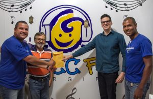 Descrição da imagem. Foto horizontal de quatro homens adultos de pé. dois a esquerda e dois a direita da imagem, sorridentes, olhando para a frente. Um homem do lado direito e um do lado esquerdo seguram um envelope ao centro da imagem. Ao fundo, parede branca com logomarca do Gacc e notas musicais. Fim da descrição.
