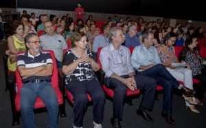 Descrição da imagem. Foto horizontal de uma plateia lotada de pessoas sentadas e apenas um rapaz em pé no fundo. As cadeiras são acolchoadas de cor vermelha, o piso é de carpete cinza e o ambiente tem pouca iluminação. Fim da descrição.