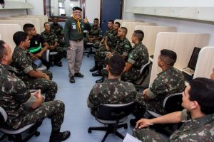 Descrição da imagem. Foto horizontal de um grupo de 15 jovens vestidos em fardamento militar, todos sentados num laboratório de informática, formando duas filas, voltados para o fundo da sala, onde aparece de pé um homem, sorridente, olhando para a frente, fazendo sinal de positivo com a mão esquerda, vestido em calça beige, camisa verde escuro, lenço amarelo no pescoço e boina. Fim da descrição.