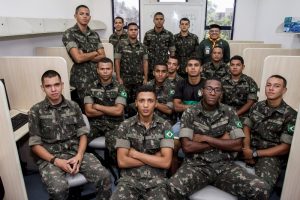 Descrição da imagem. Foto horizontal de um grupo de 15 jovens vestidos em farda militar e um homem com camisa verde escuro e lenço amarelo no pescoço, todos no meio de um laboratório de informática. Os jovens à frente estão sentados, e os do fundo da sala, estão de pé. Todos olham para a frente. Fim da descrição.