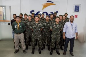 Descrição da imagem. Foto horizontal de um grupo de jovens de pé, vestidos com fardamento do exército e as mãos para trás do corpo, formando quatro alinhamentos, sendo que no primeiro, há apenas dois jovens ao centro e na extremidade esquerda, um homem de bota, calça beige, camisa verde escuro, lenço amarelo no pescoço e boina, também com os braços para trás do corpo, e na estremidade direita, um homem moreno com sapato social, calça jeans escura, camisa social clara e crachá do Senac. Ao fundo, prede branca com grande logomarca do Senac. Fim da descrição.