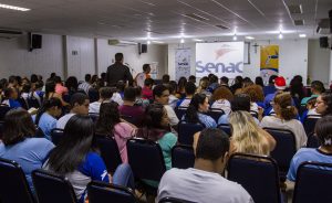 Descrição da imagem. Foto horizontal do fundo de um auditório lotado, todos sentados, exceto dois homens que caminham pelo corredor central em direção ao palco. O ambiente tem paredes e teto brancos, e ao fundo, na parede de frente para o público, uma projeção ao centro com a logomarca do Senac, do lado esquerdo, um banner branco com logo do Senac e à esquerda, um banner amarelo com logomarca do Senac Pleno. Fim da descrição.