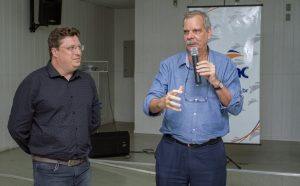 Descrição da imagem. Foto horizontal parcial de dois homens, lado a lado, porém não encostados um no outro, o da direita aparece de frente e o da esquerda de semiperfil voltado para o outro. O da direita segura um microfone com a mão esquerda próximo a boca, a qual está entreaberta, e sua mão direita está direcionada para a frente como se gesticulasse, ele é alto, tem bigode e cabelos grizalhos, óculos de grau pendurados por cordão no pescoço, usa camisa social azul claro e calça azul escuro. O homem da esquerda, está com os braços voltados para trás, tem cabelos ondulados castanhos, usa óculos de grau, olha para o homem da direita com um leve sorriso, veste camisa social preta e calça jeans. O ambiente é fechado com paredes brancas e ao fundo um banner com logomarca do Senac. Fim da descrição.