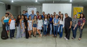 Descrição da imagem. Foto horizontal de um grupo de pessoas jovens, de pé, lado a lado, voltadas para a frente e sorridentes. Elas estão posicionadas em uma fila à frente e outra ao fundo, um degrau acima. O ambiente é fechado, com paredes e teto brancos, ao fundo, três banners, o primeiro de cor branca, o segundo, ao centro e maior também de cor branca, e um terceiro de cor amarela, onde está visível o catavento símbolo do Senac Pleno. Fim da descrição.